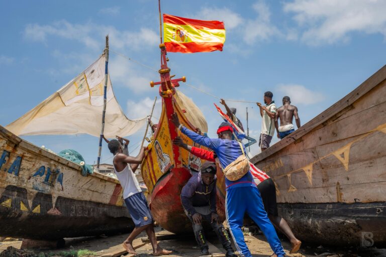 Group of men pushing colorful fishing boats on the shore with the Spanish flag flying high.